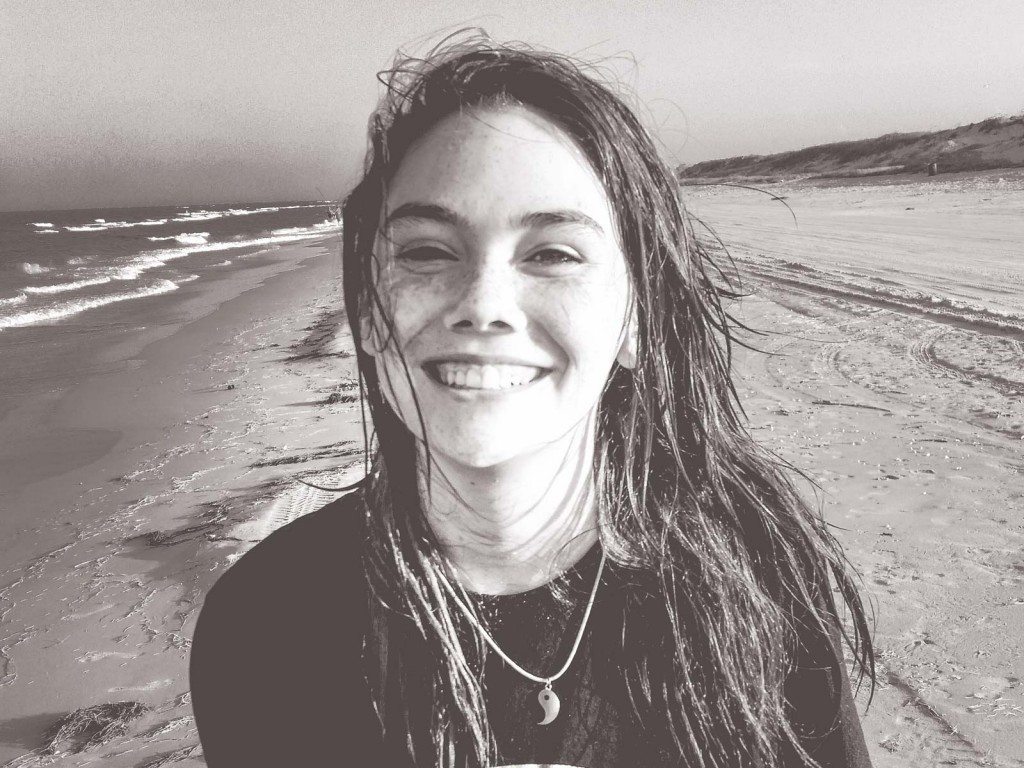 A beautiful teenage girl smiles at the camera while standing on the beach.